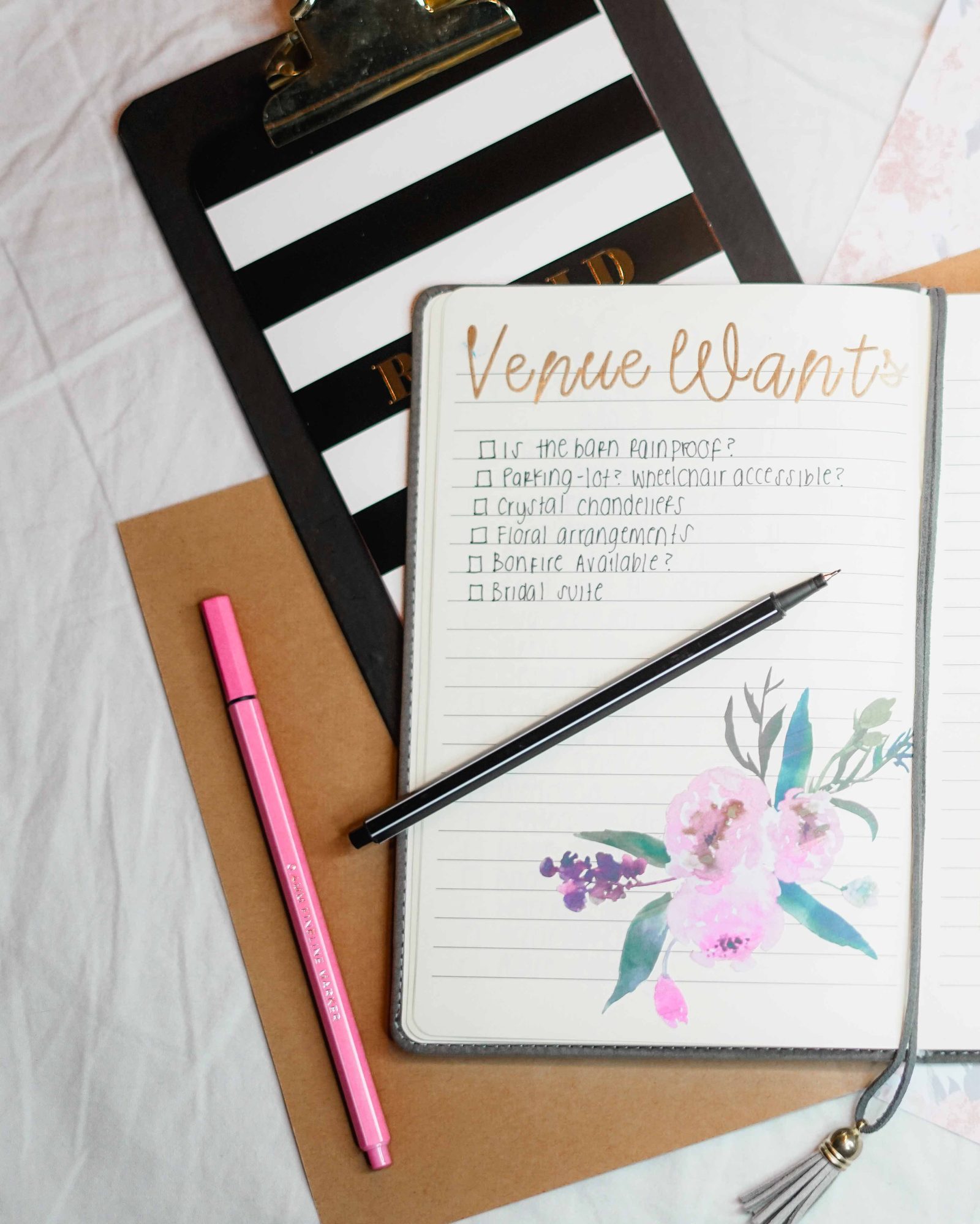 Wedding planning notebook open to a venue checklist, with a clipboard and florals
